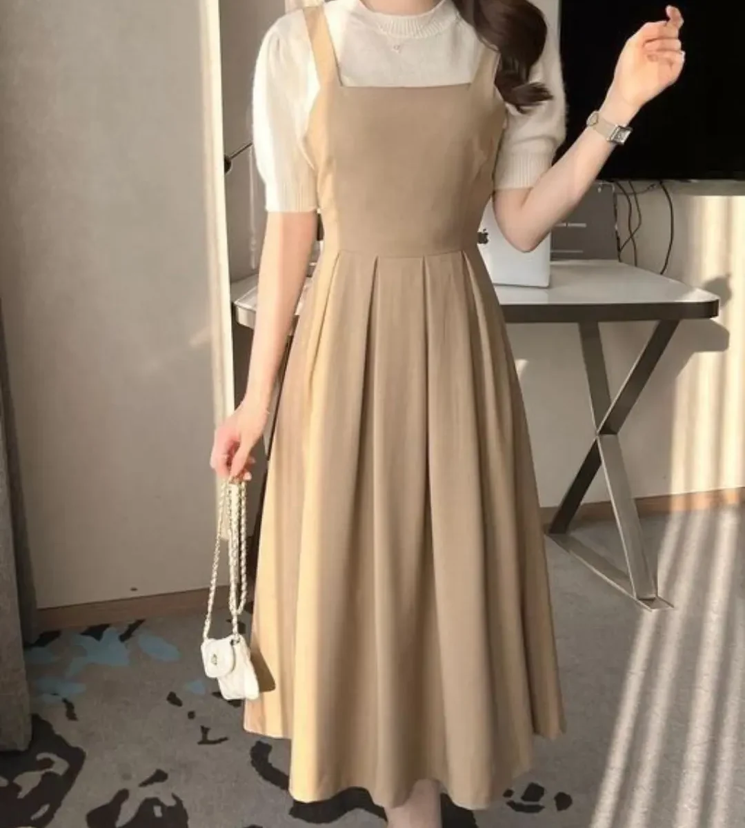 Beige sleeveless midi pinafore dress layered over a white short-sleeve top worn by a woman indoors